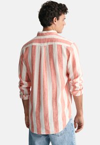 Pánská košile na knoflíky v pruhovaném provedení korálové a bílé barvy z lnu. Má dlouhé rukávy a detail na zadním díle. Volný střih s ohrnutými manžetami.