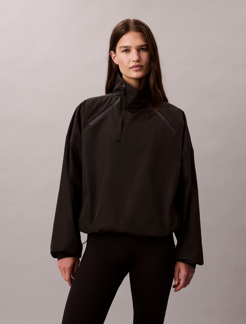 Black pullover jacket with a high collar, long sleeves, and a relaxed fit. Features a zippered front pocket and elastic cuffs.