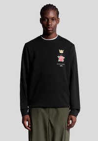 Schwarzer Sweatshirt mit geripptem Ausschnitt, der ein farbiges Grafikdesign mit einer Krone und einer Rose sowie den Text "Lyle & Scott 1874" zeigt. Das Modell trägt khakifarbene Hosen.