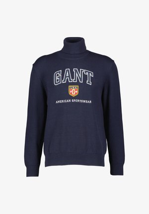 Marineblå høyhalset genser laget av teksturert stoff, med stor hvit "GANT"-logo og skjoldemblem på brystet.