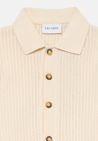 Beige knitted polo shirt with a textured pattern, featuring six round brown buttons and a classic collar. Label reads "LES DEUX."
