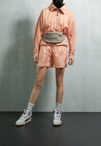 Světlý broskvový větrový kabát s límcem, doplněný o šortky ve stejné barvě. Bílé tenisky s červenými akcenty a šedý bederní batoh dokončují outfit.