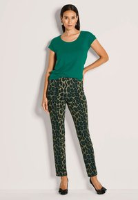 Vrouw staat met haar hand op de heupen, draagt een groene korte mouw top, een slim fit broek met luipaardprint, zwarte hakken met strikdetail, en gouden oorbellen.