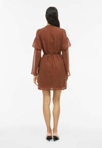 Robe marron à pois en tissu transparent, avec des manches longues à volants et une taille ceinturée, arrivant à mi-cuisse. La vue de dos montre une coupe décontractée.