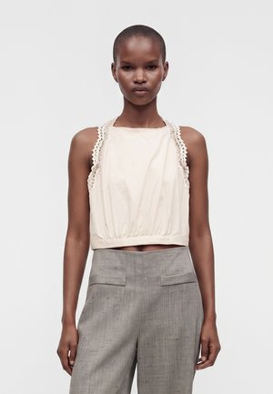 Femme aux cheveux courts portant un haut crème sans manches avec bordure en dentelle et un pantalon gris taille haute sur un fond uni.