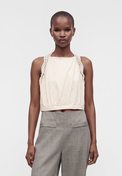 Femme aux cheveux courts portant un haut crème sans manches avec bordure en dentelle et un pantalon gris taille haute sur un fond uni.