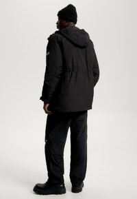 Parka negra aislante con capucha, cinta elástica en la cintura y parche del logo en la manga; lleva pantalones y botas negras.