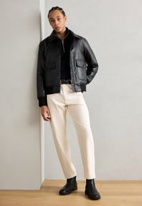 Cazadora bomber de cuero negra con dos bolsillos frontales, combinada con un suéter negro de cuello alto y pantalones color crema, terminada con botas negras hasta el tobillo.