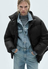 Sort puffer-jakke med høj krave, båret over en lys denimskjorte. Modellen har også en lys blå jeans på, hvilket viser et slankt, lagdelt look.