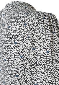 Weißer Stoff mit einem wiederholten Muster aus schwarzen und blauen Herzen. Hat eine weiche Textur und ein strukturiertes Design mit leichten Raffung an der Schulter.