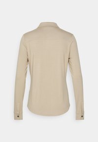 Chemise beige à manches longues avec col et poignets boutonnés, vue de dos sur un fond clair uni.