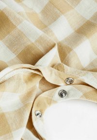 Baumwollstoff mit einem beige-weißen karierten Muster, ausgestattet mit Metall-Druckknöpfen und weichem, strukturiertem Material.