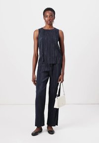 Navy pleated sleeveless top paired with matching pleated trousers, complemented by a small white handbag and flat black shoes.