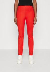 Pantaloni stretch rossi, aderenti, con una texture liscia, caratterizzati da una vita alta e gambe affusolate, abbinati a sneakers bianche.