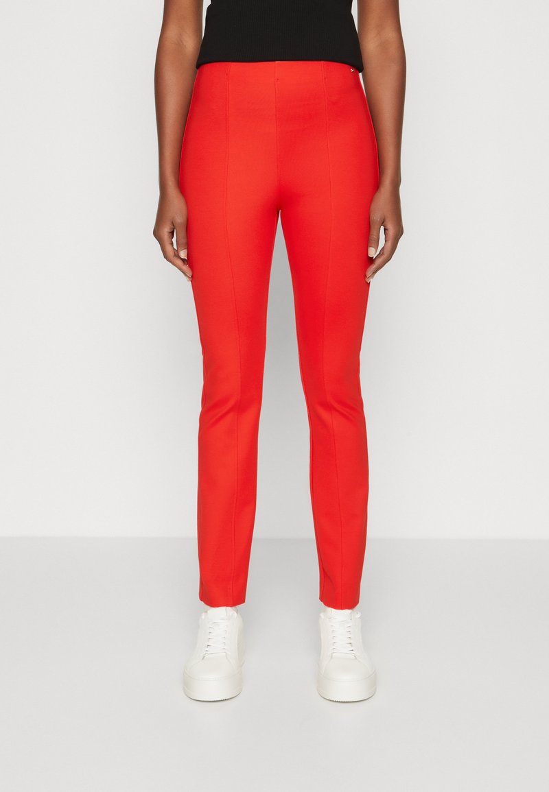 Pantaloni stretch rossi, aderenti, con una texture liscia, caratterizzati da una vita alta e gambe affusolate, abbinati a sneakers bianche.
