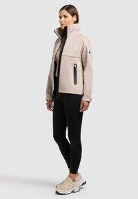 Femme portant une veste beige à col montant avec une fermeture éclair noire, des leggings noirs et des baskets beiges, debout devant un fond gris clair uni.