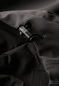 Zwarte stof met een ruitpatroon, voorzien van een zwarte koordlock met de tekst "SUPER DRY®" en een gladde, licht reflecterende textuur.