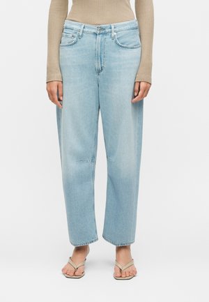 Ανοιχτό μπλε τζιν ψηλόμεσο με ίσια μπατζάκια, φοράει με μπεζ μακρυμάνικη ριμπ μπλούζα και μπεζ σανδάλια με τετράγωνη μύτη σε ανοιχτό φόντο.