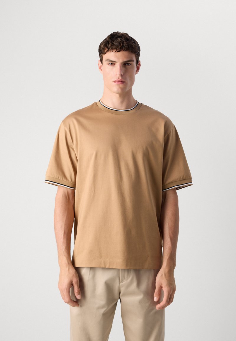 Joven vistiendo una camiseta de manga corta marrón con ribetes en negro y blanco en el cuello y las mangas, combinada con pantalones beige, de pie contra un fondo blanco.