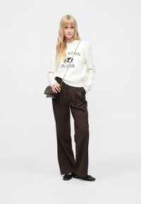 Balta džemperis ar uzrakstu "Mountain Ski Club", brūnas plaša stila bikses, melni apavi un neliela melna plecu soma ar ķēdes siksnu.