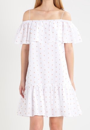 Biała sukienka z odkrytymi ramionami, cienkimi ramiączkami i falbanami, z wzorem małych beżowych kropek, noszona przez stojącą osobę.