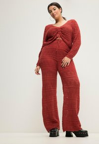 Set di maglia rosso composto da due pezzi, con una maglietta a maniche lunghe con dettaglio a fiocco e pantaloni a gamba larga. Il motivo include designs e texture a lavorazione aperta. Tacchi a piattaforma neri.