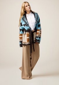 Cárdigan de punto con capucha y cinturón en un patrón multicolor, que presenta diseños geométricos en azul, marrón y crema, combinado con pantalones beige de pierna ancha.