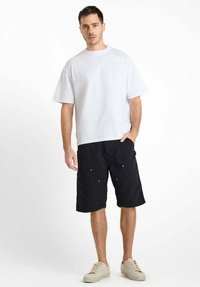 Witte korte mouwen t-shirt gecombineerd met zwarte cargo shorts tot de knie met voorkeuzes en lichte hardware accenten. Beigekleurige sneakers.