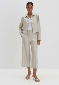 Beige linnen cropped jasje met knoopsluiting en twee voorzakken, gecombineerd met bijpassende wijde broek, aangevuld met metallic sandalen.