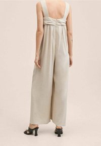 Jumpsuit en lin beige avec des jambes larges, de larges bretelles et un détail froncé dans le dos. Porté avec des sandales à talons noires.