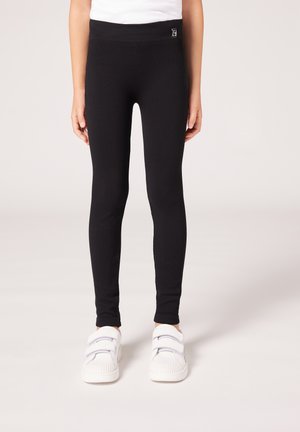 Svarta ribbade leggings med ett brett midjeband och en liten etikett. Bärs med vita velcro-sneakers med texturerade sulor.