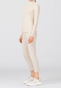 Kvinna klädd i en krämfärgad ribbat polotröja, avkortade beige byxor och vita klumpiga sneakers, som står mot en ljus, enfärgad bakgrund.