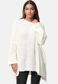 Übergroßer weißer Strickpullover mit Rippkragen und -bündchen, der ein strukturiertes Muster und seitliche Schlitze aufweist. Kombiniert mit schwarzen Jeans.