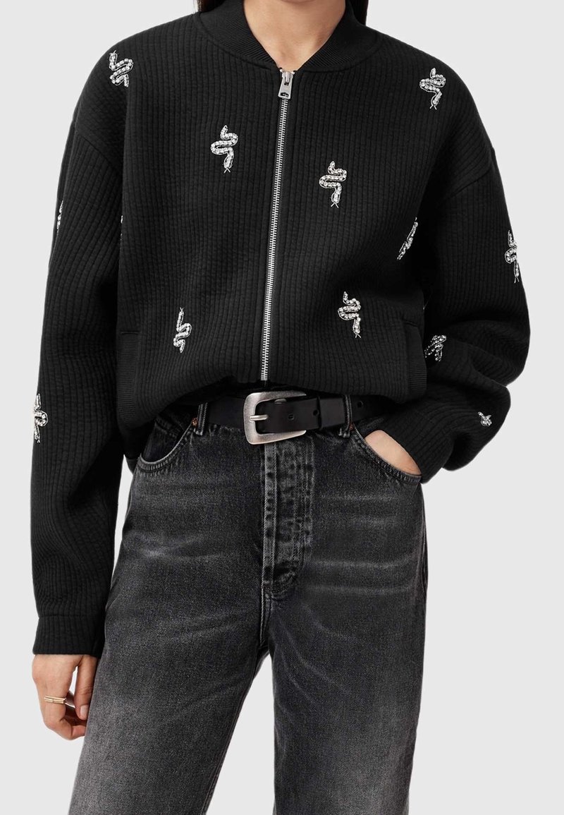 Femme portant un pull noir côtelé zippé avec une broderie de serpent blanc, associé à un jean noir délavé et une ceinture noire.