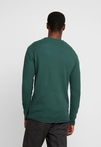 Zielony sweter o fakturze, z długimi rękawami i zaokrąglonym dekoltem, połączony z szarymi spodniami. Tkanina wydaje się przewiewna i casualowa.