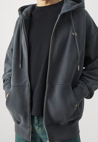 Felpa grigia con zip e finitura testurizzata, dotata di tasca frontale, cappuccio con cordino e dettaglio del logo. Indossata sopra una maglietta nera.