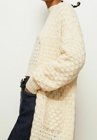 Světle béžový pletený svetr s otvorovým vzorem, pružným límcem a manžetami a přední náprsní kapsou. Nositý přes džíny.