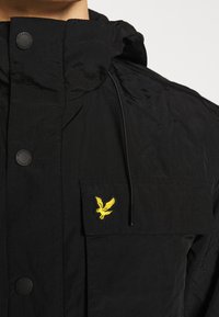 Musta takki, jossa on korkea kaulus ja läppätasku, jossa keltainen kirjailtu kotka ja mustat painonapit.