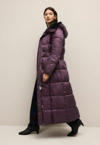 Pikk lilla puffer-kate kapuutsiga, quiltitud muster, lukuga ja kunstkarusnahast krae. Kantar tumedate teksade ja mustade pahkluusaabaste kohal.