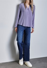 Blusa a maniche lunghe, morbida, di un leggero colore viola, con scollatura a V con bottoni, abbinata a jeans larghi in denim scuro e sneakers bianche.