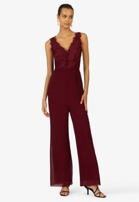 Burgunderfarbener Jumpsuit mit V-Ausschnitt und aufwendigen Spitzenverzierungen am Oberteil, weit geschnittenem Bein und einer glatten, fließenden Stofftextur.