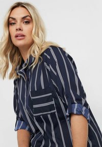 Camicia blu navy con strisce verticali bianche, chiusura con bottoni frontale, maniche arrotolabili e una tasca sul petto. Tessuto con una trama liscia.