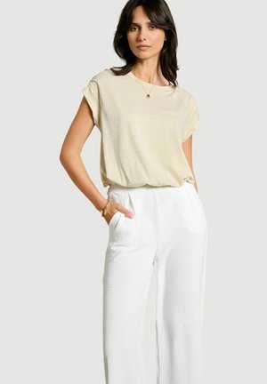 Donna con i capelli scuri che indossa una maglietta beige a maniche corte, pantaloni bianchi a vita alta, collana e braccialetto d'oro, posando con una mano nella tasca.