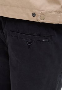 Μπεζ σακάκι με κουμπώματα με πατιλέτα φορεμένο πάνω από μαύρο παντελόνι. Το παντελόνι διαθέτει μπροστινή τσέπη με κουμπί και μικρή ετικέτα με το λογότυπο της μάρκας.