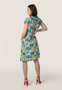Blumenkleid in Grün mit orangefarbenen und lila Mustern, kurzen Ärmeln, knielangem Saum und tailliertem Schnitt. Rote High Heels.