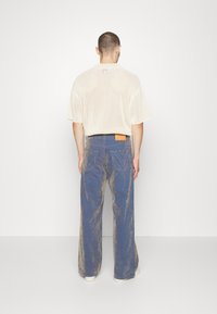 Svetle béžové síťované tričko s krátkými rukávy, spárováno s širokými modrými kalhotami s vybledlou texturou a trojúhelníkovým stehem na kapsách.