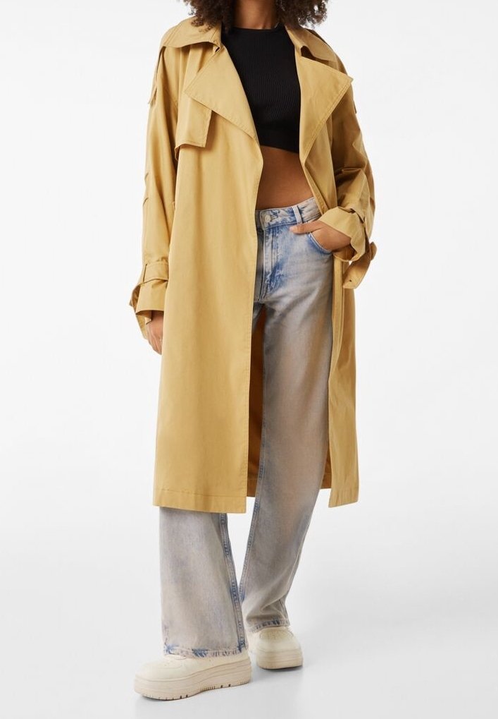 Manteau trench beige avec un col cranté, en matériau léger, poches latérales, porté sur un crop top noir et un jean en denim clair. Baskets à plateforme blanches.