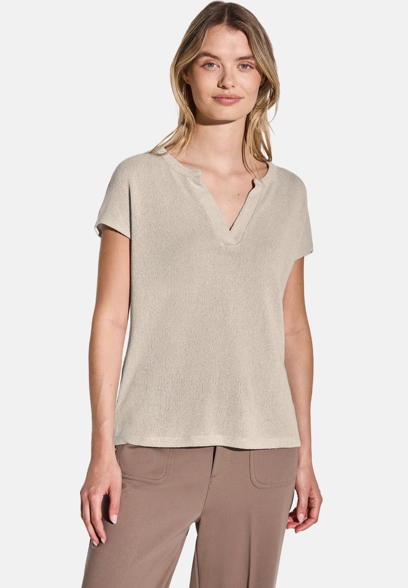 Světlý béžový top s krátkým rukávem, V-výstřihem a strukturovanou tkaninou, s uvolněným střihem a lemem, který sahá k bokům.