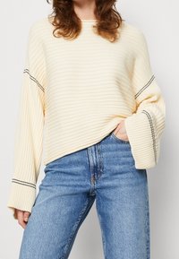 Cream ribbed sweater with loose fit, wide sleeves, and black seam detailing. Paired with blue high-waisted jeans.