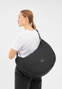 Borsa a spalla nera in tessuto morbido con forma arrotondata, tracolla regolabile e una toppa con logo visibile sulla parte anteriore. Design semplice e minimalista.